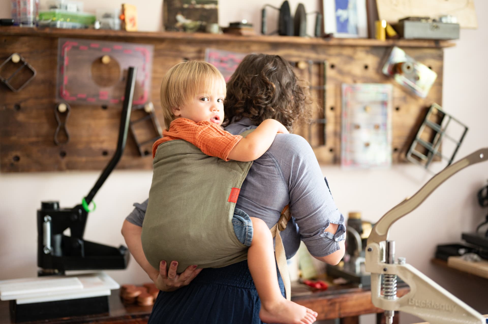 Sarah with her toddler in a carrier on her back, tool wall behind them