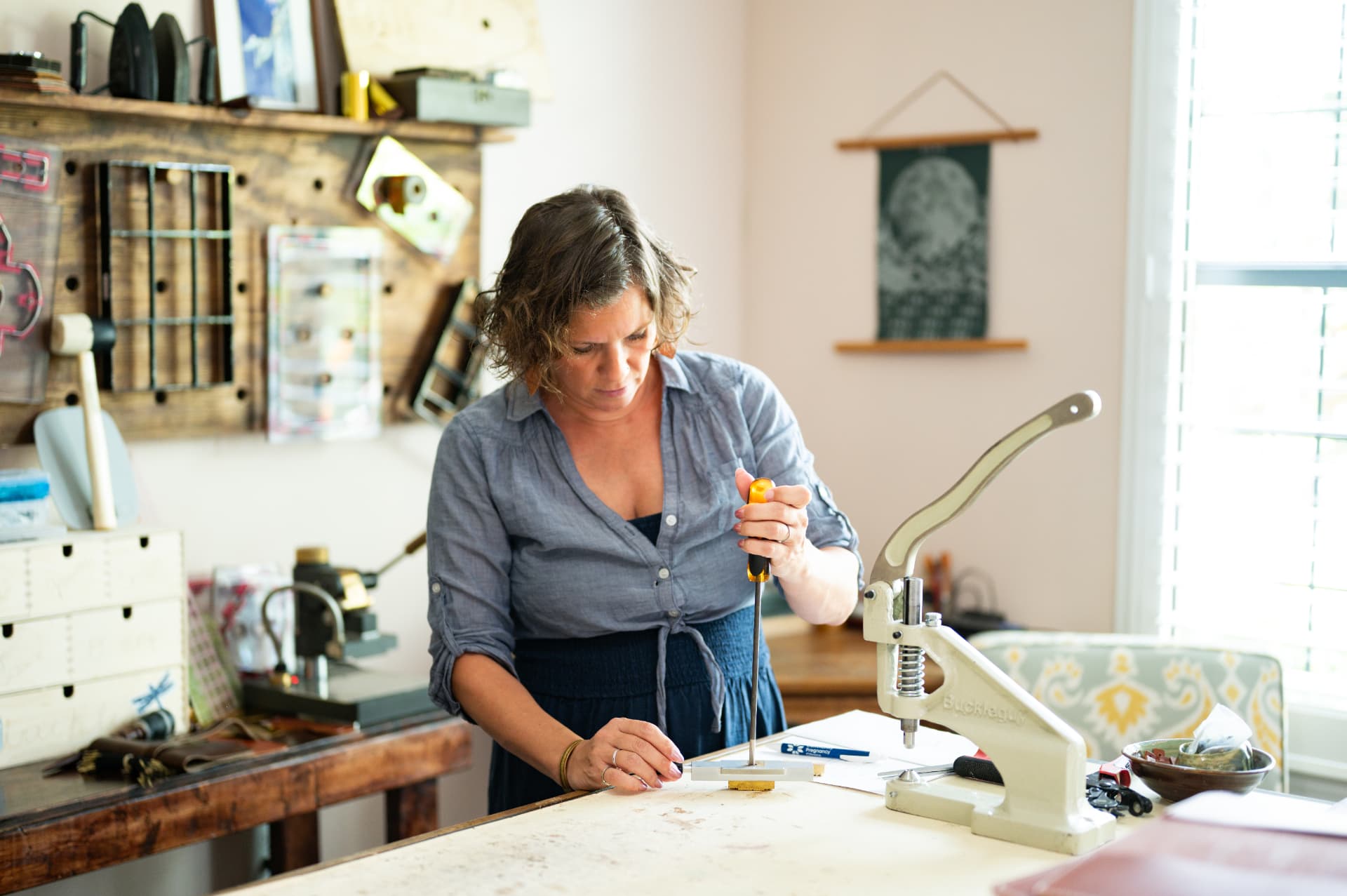 Sarah working at her bench, tool wall and window light behind her